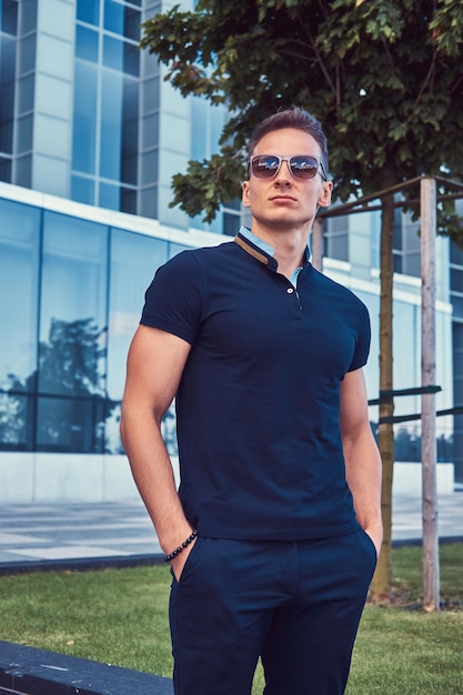 Un jeune bel homme à la mode avec une coupe de cheveux élégante en lunettes de soleil, vêtu d'un t-shirt et d'un pantalon noirs, debout dans la ville moderne contre un gratte-ciel.