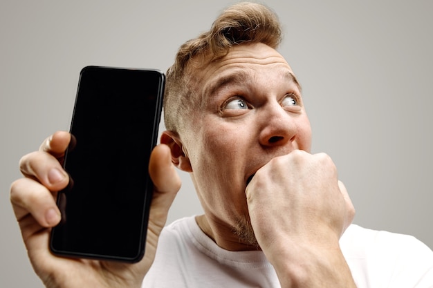 Jeune bel homme montrant l'écran du smartphone sur fond gris avec un visage surprise. Émotions humaines, concept d'expression faciale