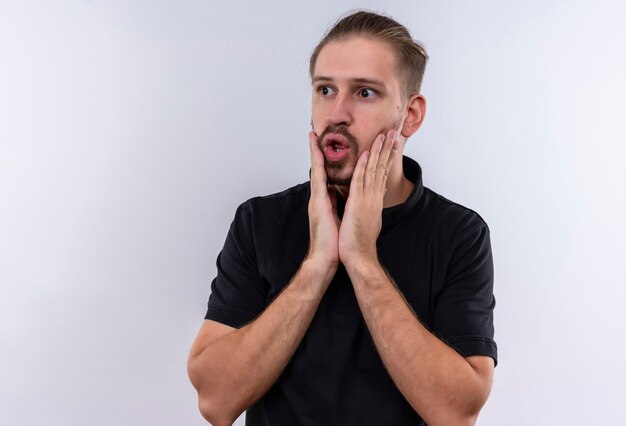 Jeune bel homme en polo noir à la surprise et étonné toucher le visage debout sur fond blanc