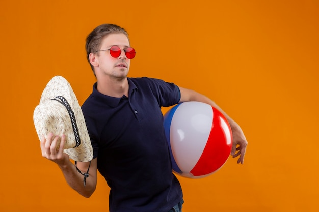 Jeune bel homme portant des lunettes de soleil rouges debout avec ballon gonflable en agitant avec chapeau de paille à la confiance sur fond orange