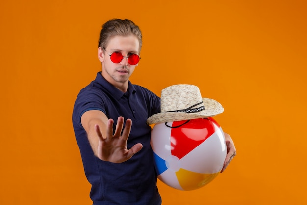 Jeune bel homme portant des lunettes de soleil rouges tenant ballon gonflable et chapeau de paille d'été debout avec la main ouverte faisant le geste d'arrêt avec un visage sérieux sur fond orange