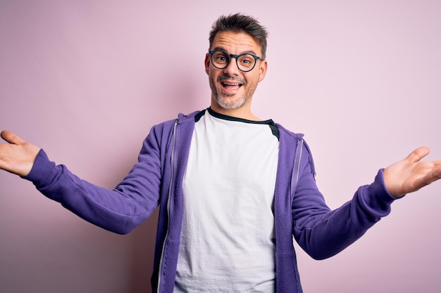 Photo gratuite jeune bel homme portant un sweat-shirt violet et des lunettes debout sur fond rose souriant joyeux offrant des mains donnant de l'aide et de l'acceptation