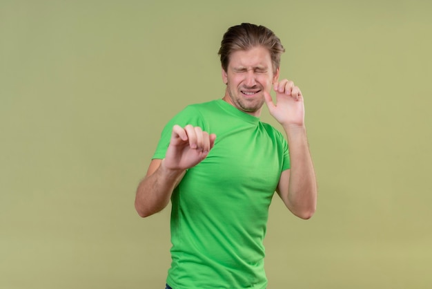 Jeune bel homme portant un t-shirt vert debout avec les yeux fermés faisant le geste de défense tenant la main debout sur le mur vert