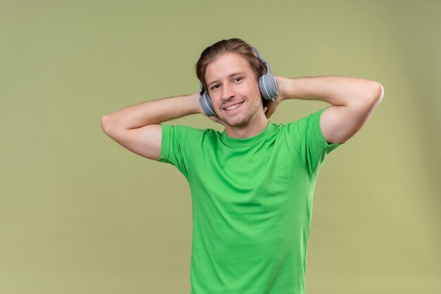 Jeune bel homme portant un t-shirt vert avec des écouteurs appréciant la musique préférée debout sur le mur vert