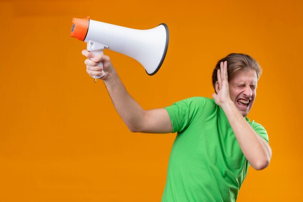 Jeune bel homme portant un t-shirt vert protégeant avec la main contre un mégaphone avec les yeux fermés agacé debout sur un mur orange