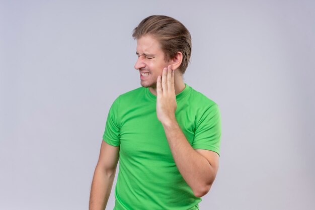 Jeune bel homme portant un t-shirt vert à la recherche de mal à toucher sa joue ayant mal aux dents