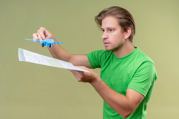 Jeune bel homme portant un t-shirt vert tenant un avion jouet et une carte avec une expression sérieuse sur le visage debout sur mur vert