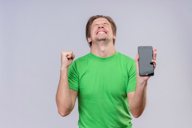 Jeune bel homme portant un t-shirt vert tenant un téléphone mobile fou heureux serrant le poing se réjouissant de son succès debout sur un mur blanc