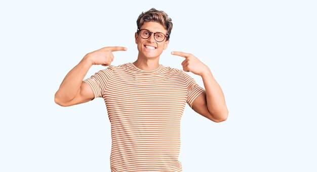 Jeune bel homme portant des vêtements décontractés et des lunettes souriant joyeux montrant et pointant avec les doigts les dents et la bouche. concept de santé dentaire.