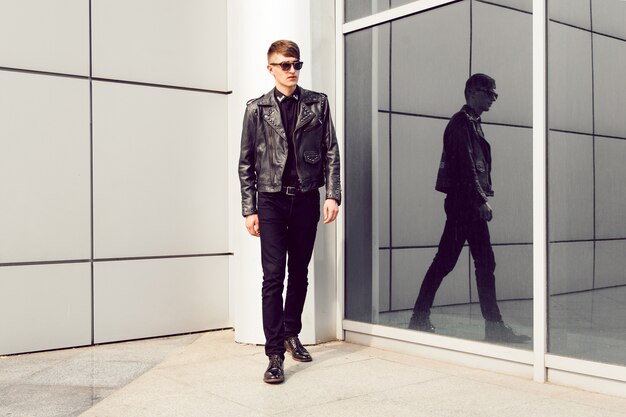 Jeune bel homme posant près du centre d'affaires moderne, vêtu d'une élégante veste en cuir à pointes, d'un jean noir et de lunettes de soleil, look brutal.