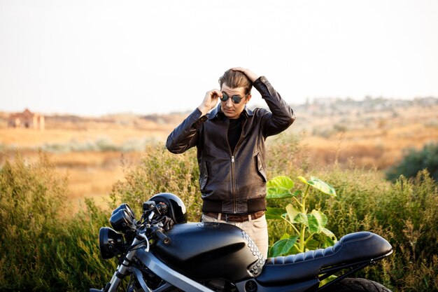 Jeune bel homme posant près de sa moto à la route de campagne.