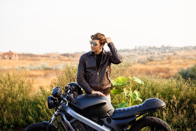 Jeune bel homme posant près de sa moto à la route de campagne.