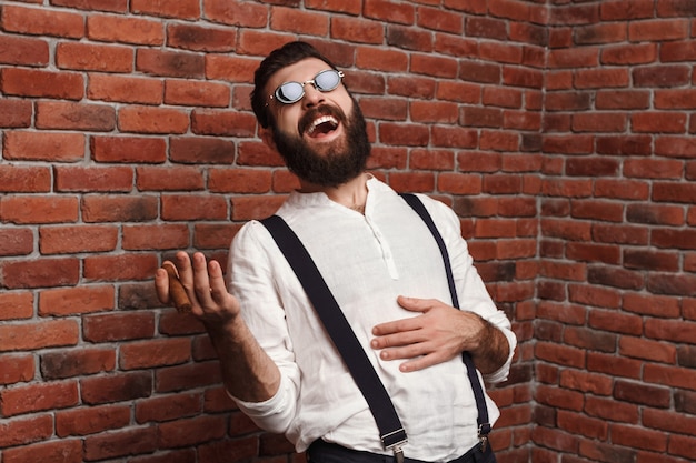 Photo gratuite jeune bel homme riant tenant un cigare sur le mur de briques.