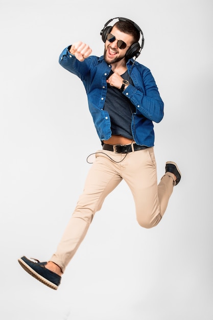 Jeune bel homme souriant heureux dansant et écoutant de la musique dans des écouteurs isolé sur fond de studio blanc, portant chemise en jean et lunettes de soleil, gagnant sautant dans le succès