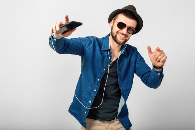 Jeune bel homme souriant heureux, écouter de la musique dans des écouteurs isolé sur fond de studio blanc, tenant un smartphone, portant une chemise en jean, un chapeau et des lunettes de soleil