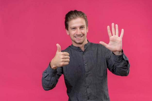 Jeune bel homme souriant montrant et pointant vers le haut avec les doigts numéro six debout sur le mur rose