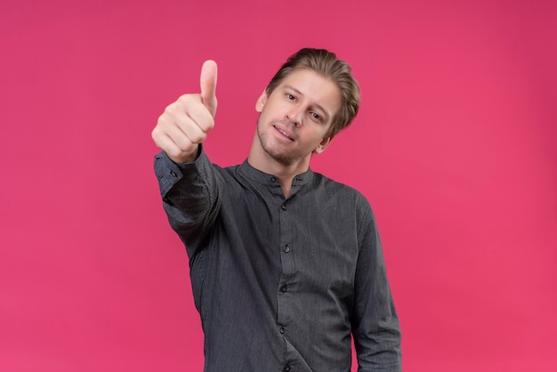 Jeune bel homme souriant sympathique montrant les pouces vers le haut debout sur un mur rose