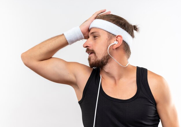 Jeune bel homme sportif confus portant un bandeau et des bracelets et des écouteurs mettant la main sur la tête en regardant le côté isolé sur le mur blanc