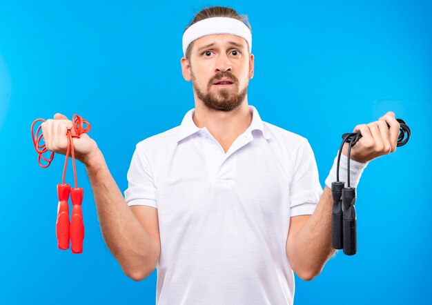 Jeune bel homme sportif confus portant un bandeau et des bracelets tenant des cordes à sauter isolées sur un mur bleu