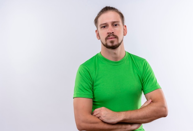 Photo gratuite jeune bel homme en t-shirt vert debout avec les bras croisés regardant la caméra avec un visage sérieux sur fond blanc