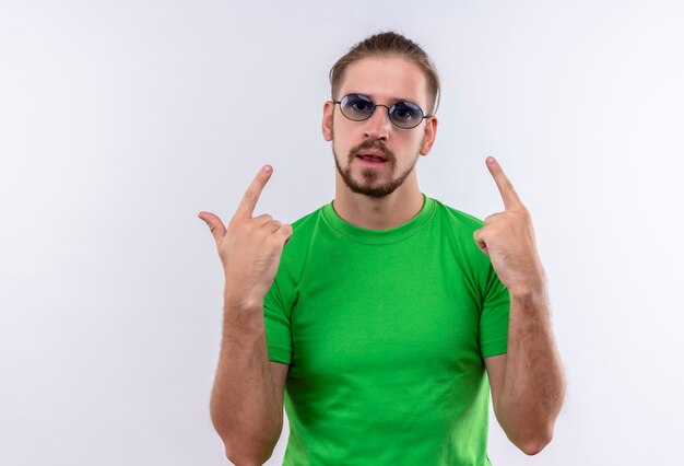 Jeune bel homme en t-shirt vert portant des lunettes pointant avec les doigts vers le haut à confiant debout sur fond blanc