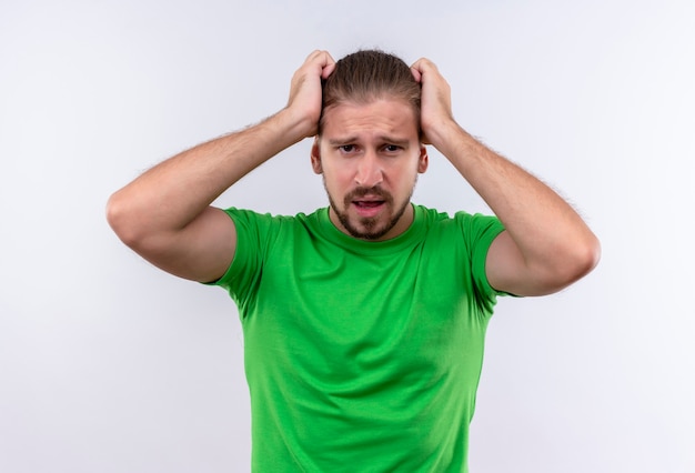 Jeune bel homme en t-shirt vert regardant la caméra déçu de toucher sa tête debout sur fond blanc