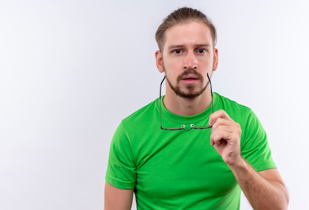 Jeune bel homme en t-shirt vert regardant la caméra avec une expression sérieuse sur le visage tenant des lunettes à la main debout sur fond blanc