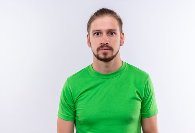 Jeune bel homme en t-shirt vert regardant la caméra surpris et perplexe debout sur fond blanc