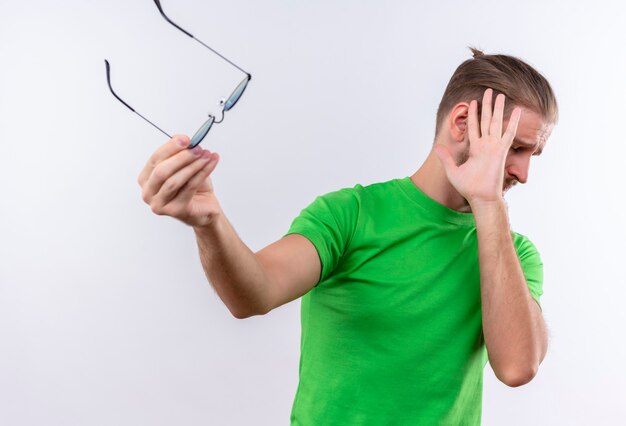 Jeune bel homme en t-shirt vert tenant des lunettes à la main faisant le geste de défense debout sur fond blanc