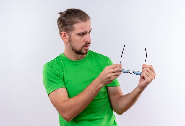 Jeune bel homme en t-shirt vert tenant des lunettes de soleil dans les mains à la recherche de côté avec une expression sérieuse debout sur fond blanc