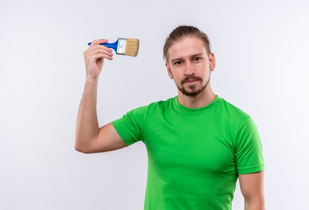 Jeune bel homme en t-shirt vert tenant un pinceau à la confiance debout sur fond blanc