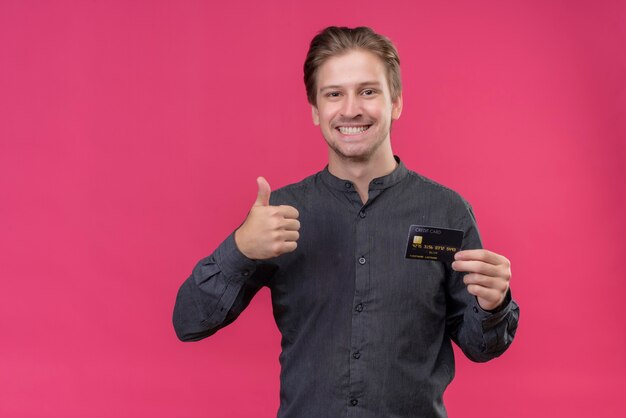 Jeune bel homme tenant une carte de crédit montrant les pouces vers le haut, souriant debout sur un mur rose