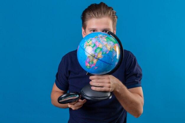Jeune bel homme tenant un globe et une loupe se cachant derrière un globe debout sur fond bleu