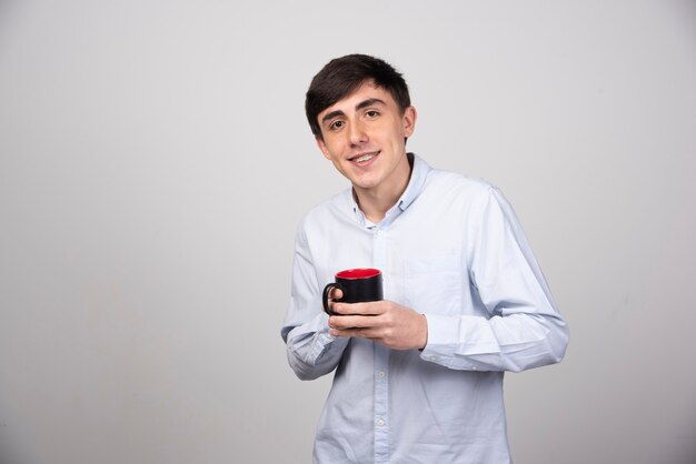 Jeune bel homme tenant une tasse de thé sur un mur gris.