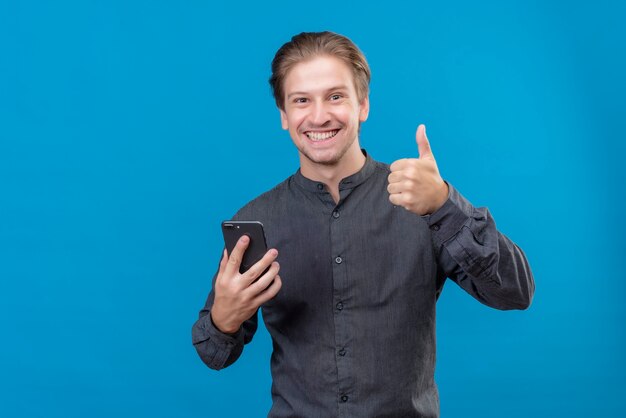 Jeune bel homme tenant un téléphone mobile montrant les pouces vers le haut souriant debout sur le mur bleu