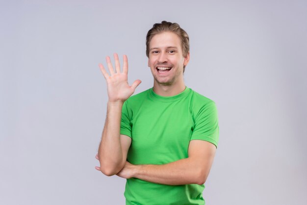 Jeune bel homme vêtu d'un t-shirt vert en agitant avec la main debout sur un mur blanc