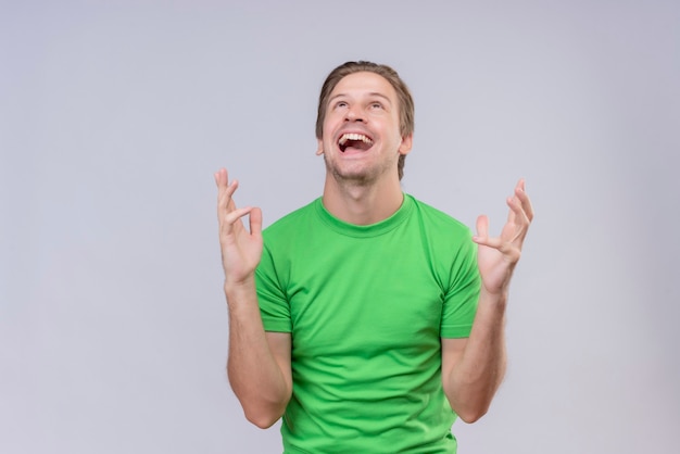 Jeune bel homme vêtu d'un t-shirt vert fou heureux avec les bras levés debout sur un mur blanc 3