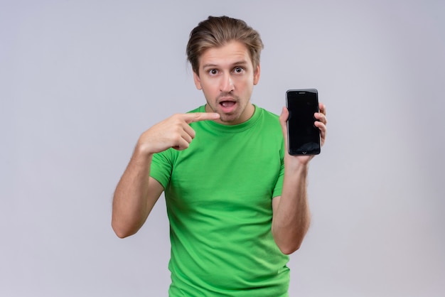 Jeune bel homme vêtu d'un t-shirt vert tenant le smartphone pointant avec le doigt dessus à la surprise et confus debout sur un mur blanc