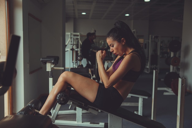 Jeune belle athlète blonde sexy bodybuilder exerce dans la salle de gym dans de beaux vêtements de sport sur un fond sombre