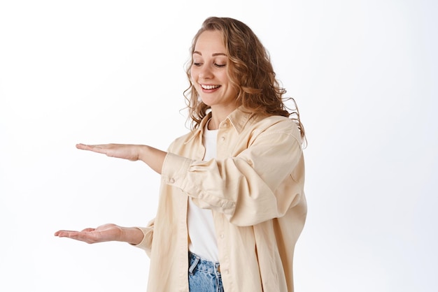 Jeune belle étudiante blonde tenant un fond vide entre les mains montrant le logo ou le produit de grande taille souriant et regardant heureux à l'article fond blanc