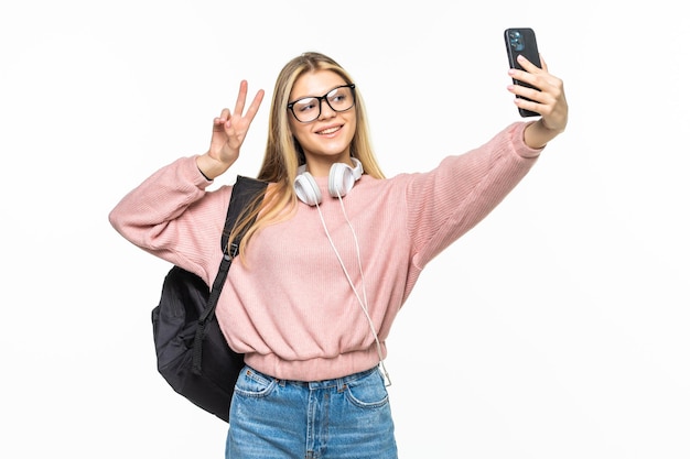 Jeune belle étudiante avec sac à dos fait selfie isolé sur mur blanc en studio