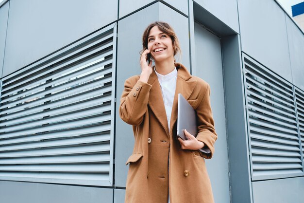 Jeune belle femme d'affaires décontractée en manteau avec ordinateur portable parlant joyeusement sur téléphone portable en plein air