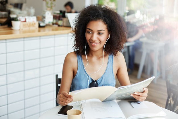 Jeune belle femme africaine étudiante dans les écouteurs assis dans un café souriant tenant le livre. Éducation et apprentissage.