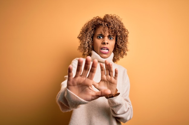 Photo gratuite jeune belle femme afro-américaine portant un pull à col roulé sur fond jaune effrayée et terrifiée par l'expression de la peur arrêter le geste avec les mains criant sous le choc concept de panique