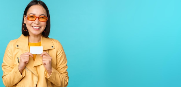 Jeune belle femme asiatique montrant une carte de crédit en souriant en choisissant une banque debout sur fond bleu