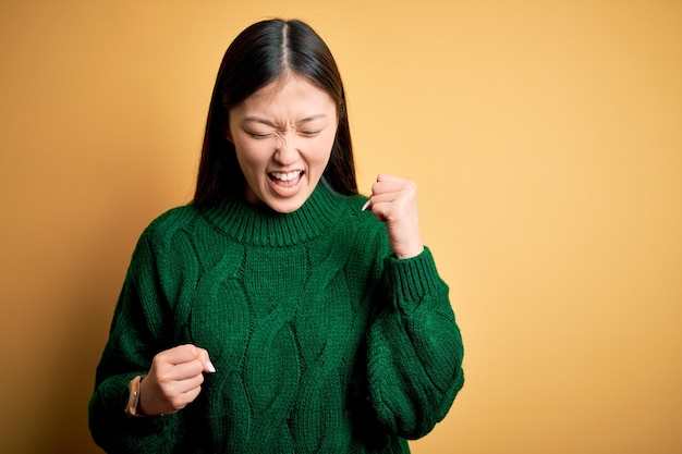 Photo gratuite jeune belle femme asiatique portant un pull d'hiver vert sur fond isolé jaune célébrant la surprise et l'émerveillement du succès avec les bras levés et les yeux fermés concept gagnant