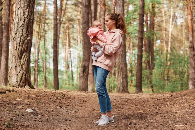 Jeune belle femme aux cheveux bruns et queue de cheval tenant une petite fille dans les mains, marchant ensemble dans un parc ou une forêt