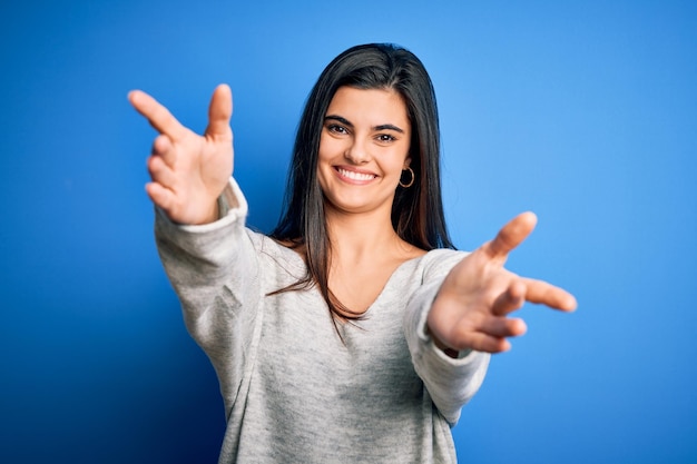 Photo gratuite jeune belle femme brune portant un pull décontracté debout sur fond bleu regardant la caméra en souriant à bras ouverts pour un câlin expression joyeuse embrassant le bonheur