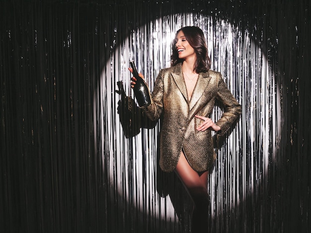 Jeune belle femme brune souriante en veste dorée de soirée à la modeSexy femme insouciante posant près du mur de guirlandes brillantes argentées en studio