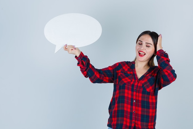 Jeune belle femme en chemise décontractée, jeans tenant une bulle de dialogue tout en gardant la main sur la tête et l'air satisfait, vue de face.